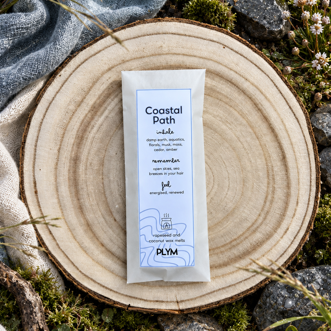 A single Coastal Path wax melt is centred on a round natural wood slice, surrounded by moss, rocks, dried grass, and a textured blue-gray cloth in an outdoor styled setting.